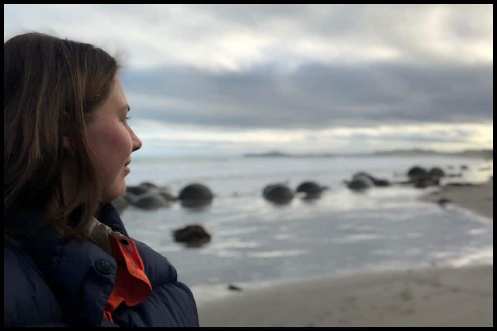 Photo shows the side profile of a young woman's face as she looks behind her at the Moeraki Boulders at low tide.