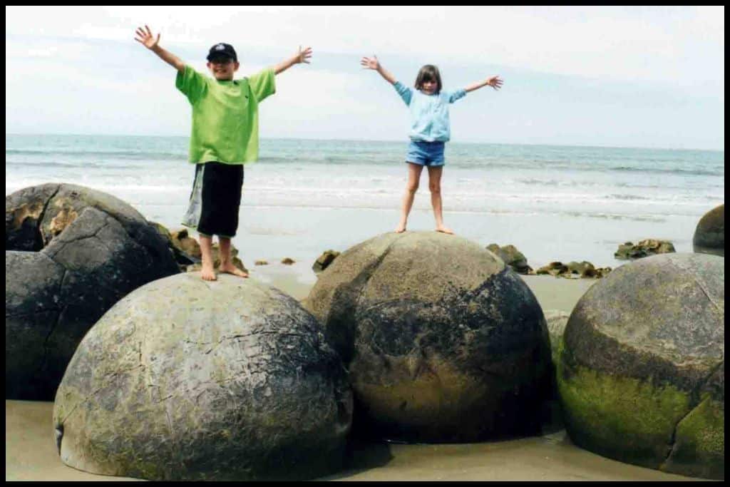 Two children raise their arms up above them, each standing on a famous Moeraki Boulder.