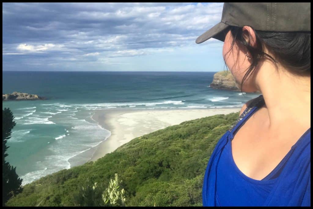 Photo shows the side profile of a young woman looking down onto an Otago beach with beautifully coloured sand and water.