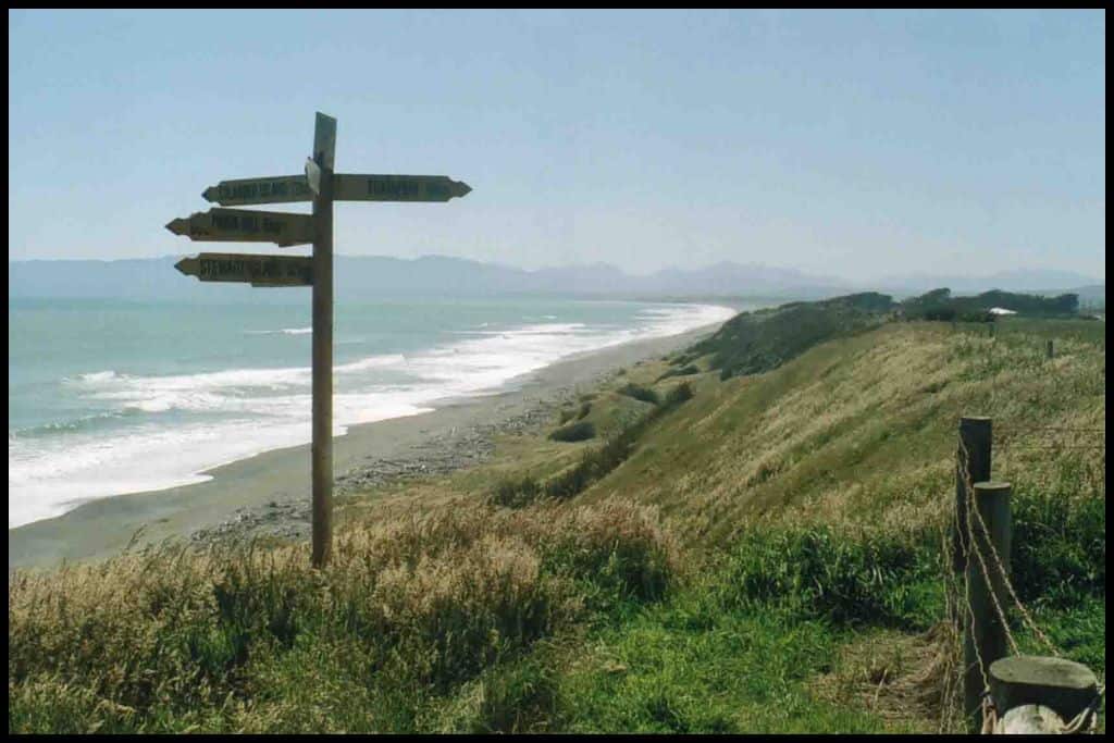 A signpost points several directions, pitched into the grassy bank above the shoreline of a Southland beach.