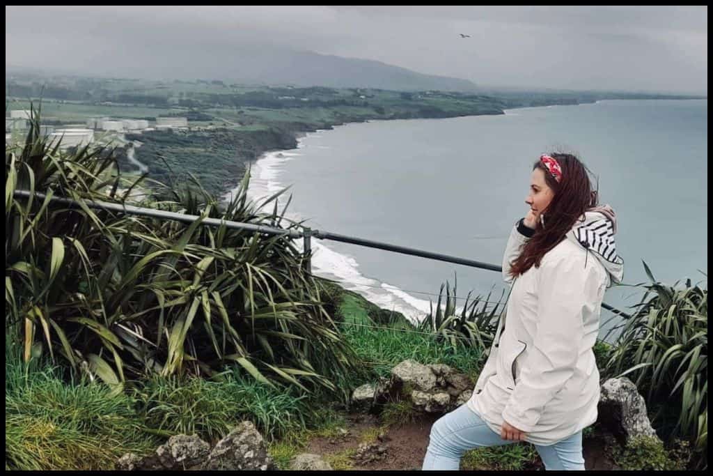 Side profile of young woman posing for the camera atop Paritutu Rock on a gloomy day with Back Beach visible below.
