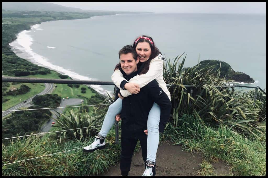 Young couple piggybacking each other laugh for the camera atop Paritutu Rock with Back Beach on a gloomy day visible behind.