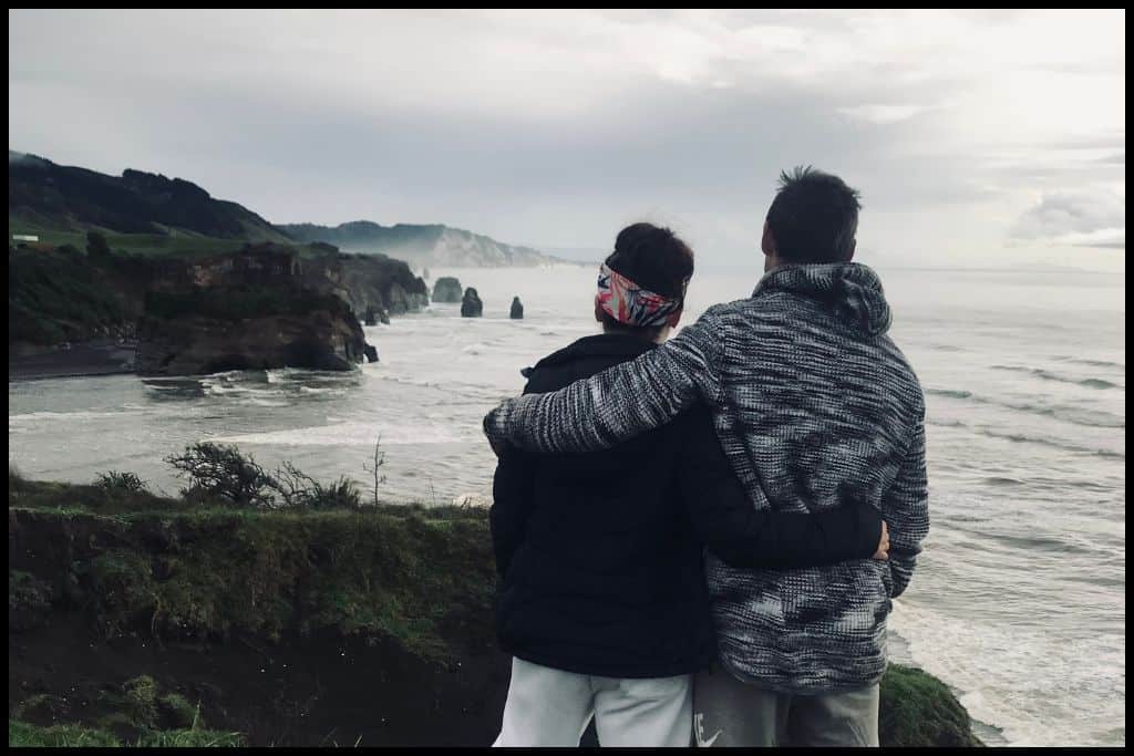 Tongaporutu Beach on a gloomy day, with the Three Sisters and Elephant Rock visible in the water, and a couple with their arms around each other in the foreground, photographed from behind.