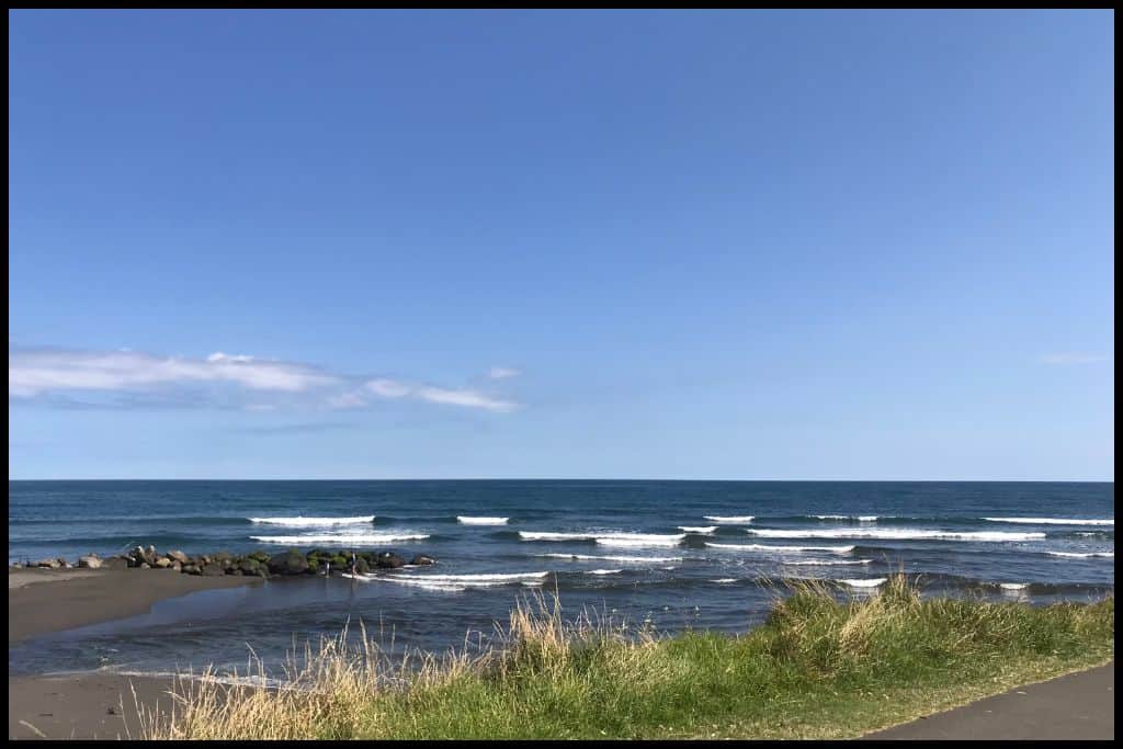 Rolling waves and a flat horizon, black sand and grass in the foreground.