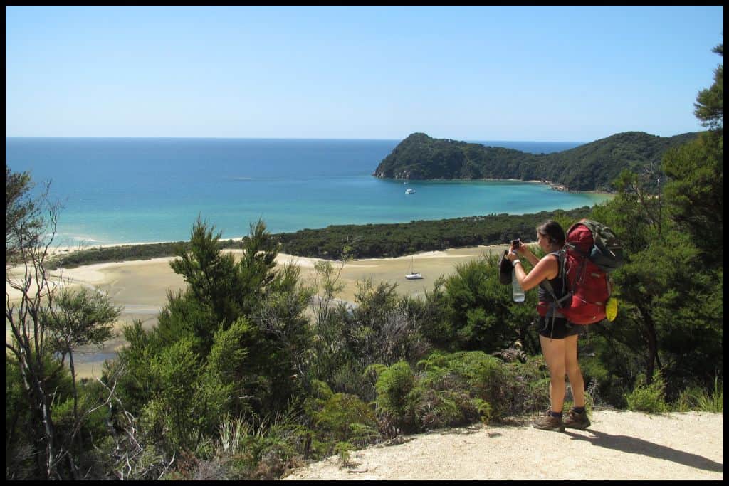 A solo hiker stops to take a photo from the side of the track of the bay below.