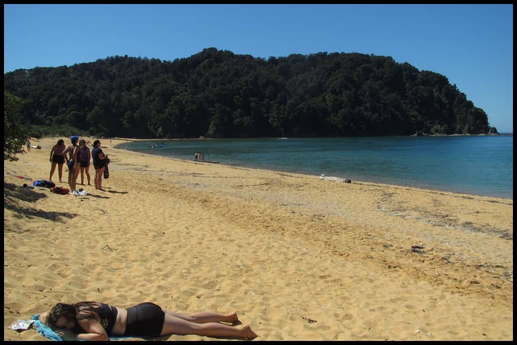 A young woman lies face down on the golden sands of Totaranui Beach, sunbathing.