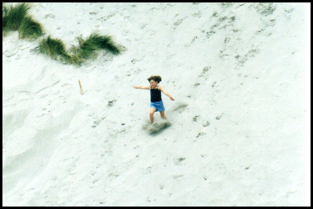 A young girl's feet are clouded in flying sand as she herself flies down a large white sand dune.