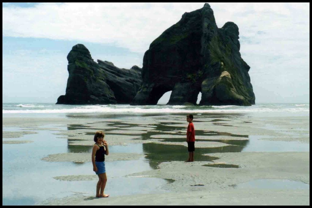 Two children stand on the sand at low tide in front of the Archway Islands that make Wharariki Beach so famous.