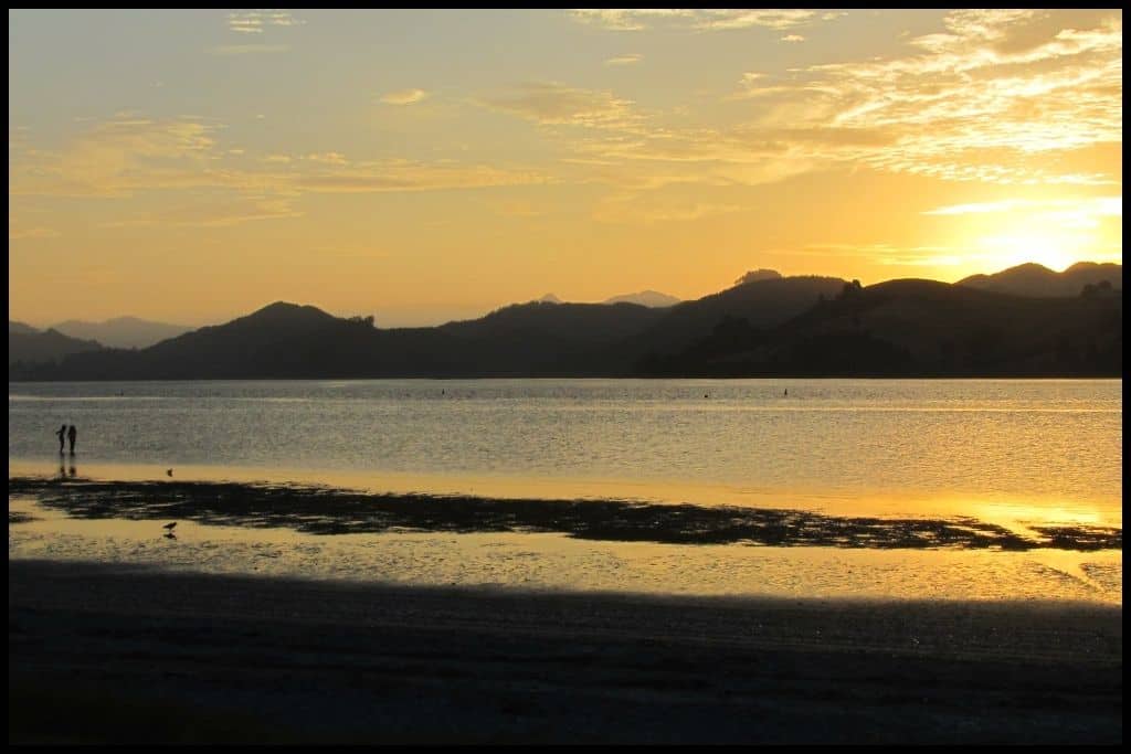 Sunset streaks across the water of the Tairua Harbour while the headlands are silhouetted against the horizon.