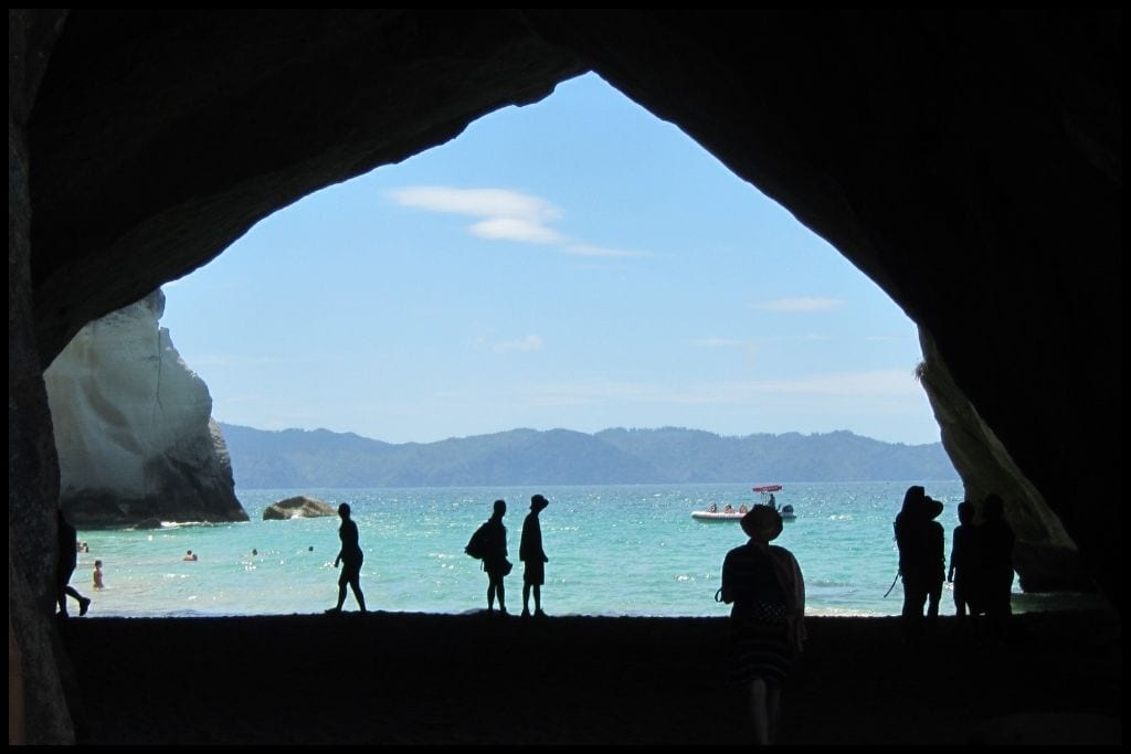 People are silhouetted against the dazzling blue of Cathedral Cove beyond the cathedral rock formation that you can walk through.