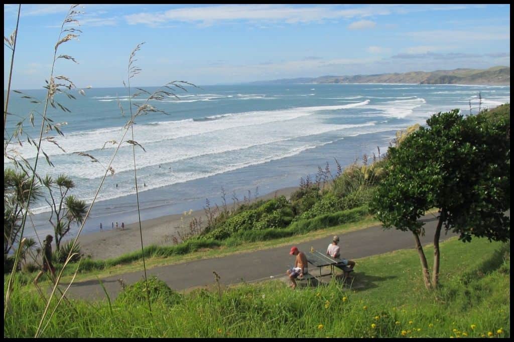 Two people sit on a bench, photographed from higher up the hill, overlooking the expanse of Ngarunui Beach below.