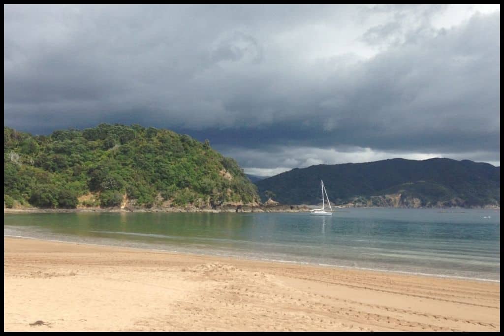 Stormclouds gather over little, picturesque, Little Waikawau Bay with its green water, golden sand and sole yacht.