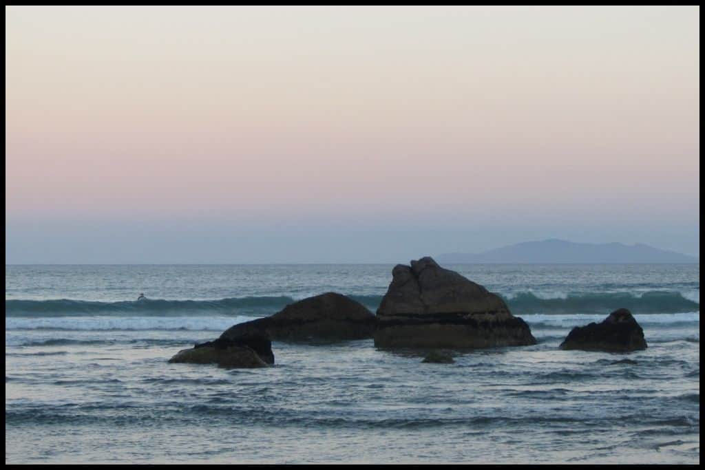 Whangamata Beach at sunset - pastel colours paint the sky. The foreground is rocks and surf.