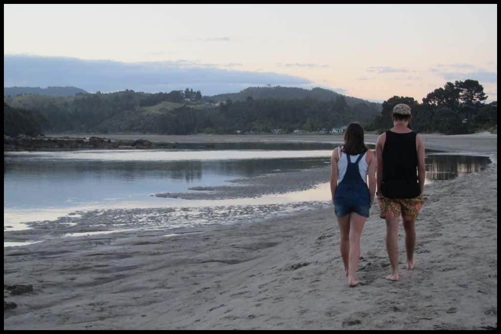 Two teens walk away from the camera down a sandy stretch of Whangamata Beach.