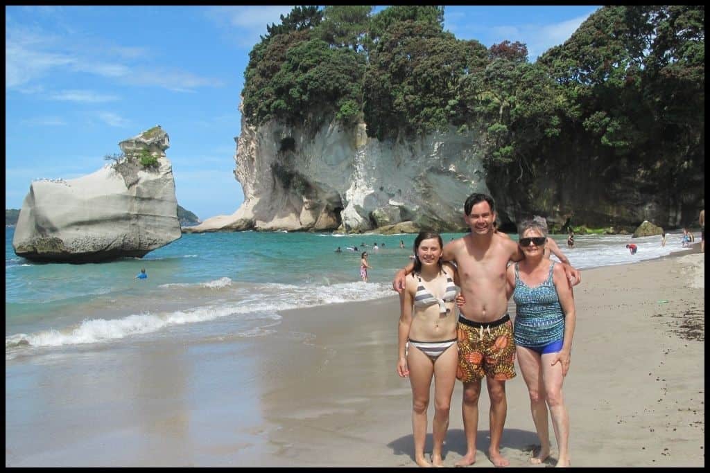 Two teens and their mum stand on the beach at Cathedral Cove with Smiling Sphinx Rock and the water behind.