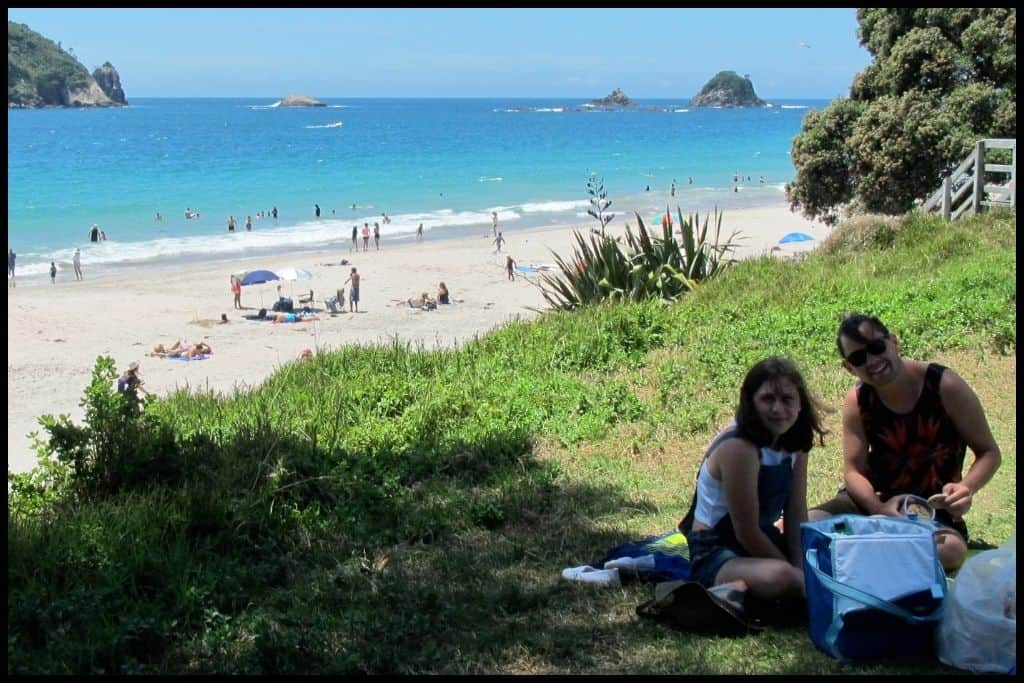 Two teens sit in the shade having a picnic with the surf of Hahei Beach pictured behind.