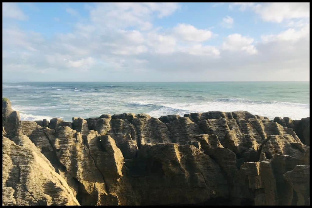 Late afternoon sun illuminates the Punakaiki Pancake Rocks, with ocean waves behind.